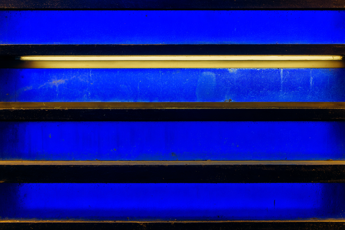 Four horizontal dark glass panels stacked with hairline gaps between them, each panel catching a different density of cobalt-blue volumetric light — bottom panel barely lit